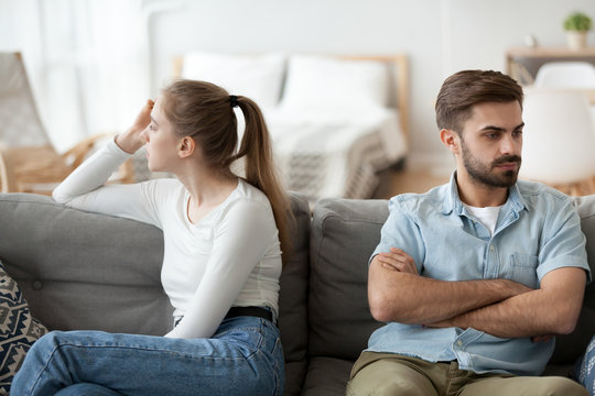 Stubborn couple sit on couch back to back avoid talking after fight, offended man and woman separated on sofa ignoring one another, husband and wife not looking in eyes. Family conflict concept