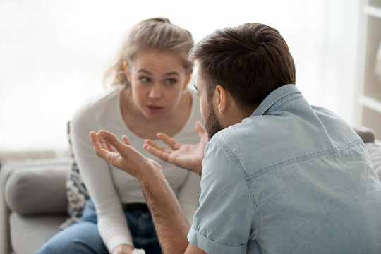 Millennial couple arguing sitting on couch at home, emotional husband gesturing proving his point of view to stubborn wife, man and woman dispute having fight or disagreement. Family conflict concept
