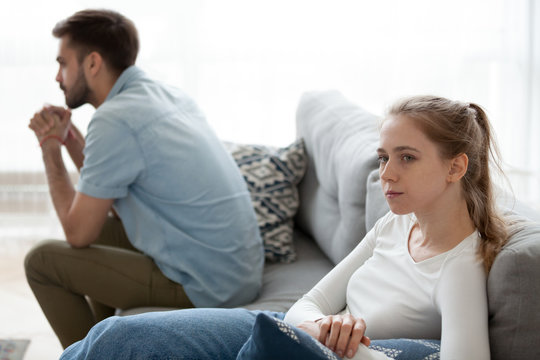 Offended husband and wife sit separately on different sides of couch avoiding talking or looking at each other, angry couple ignore one another after fight or dispute, spouses breaking up or split