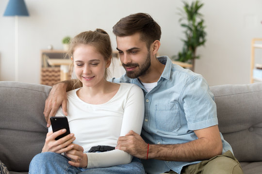 Happy young woman holding smartphone watching something with beloved, excited millennial couple relax on couch using cellphone, smiling boyfriend and girlfriend hug on sofa looking on mobile
