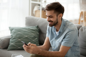 Happy man relax on couch at home chatting with friends on smartphone, excited millennial male writing message on cell to girlfriend, smiling guy using mobile phone texting or playing app games