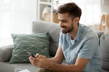 Happy man relax on couch at home chatting with friends on smartphone, excited millennial male...