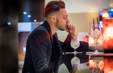 Handsome young man drinking cocktail at bar counter, wearing business suit and looking at camera