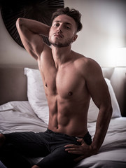 Shirtless sexy male model lying alone on his bed in his bedroom, looking at camera with a seductive attitude