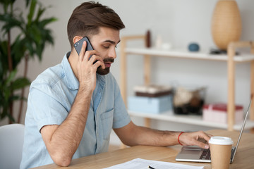 Busy millennial man sit on office table working on laptop talking on smartphone, thoughtful male use computer having conversation on cell with client, freelancer outsource from home speaking on mobile