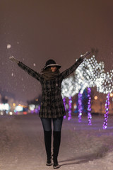 Young girl throwing snow and walking in front of trees decorated with colorful lights for christmas night, blurred in the background