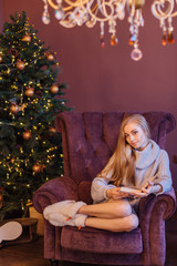 Beautiful young woman dressed in a sweater sitting on the arm chair next to the christmas tree.