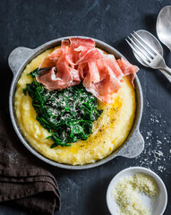Polenta with butter stewed spinach and prosciutto in a frying pan. Delicious lunch in a mediterranean style. On a dark background, top view
