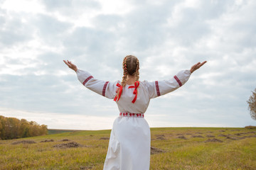 A girl in a Slavic dress with a red ribbon in her hair is standing in a field with her arms open to the sky