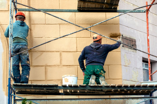 two workers in a helmet and construction clothes on a high staircase are painting with rollers, repairing the facade of the house