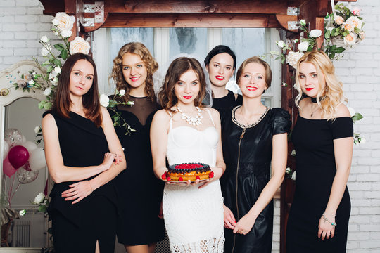 Portrait Of Beautiful Bridesmaids In Black Dresses With Attractive Bride-to-be In The Middle Holding Delicious Berry Cake. Background Is Decorated With Flowers. 