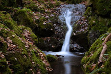 Naklejka na ściany i meble Small hidden waterfall on a hike through a forest, wonderful autumn scene with moss, small stream and blurred water flowing from the rocks. Quiet, clean and serene scenery.