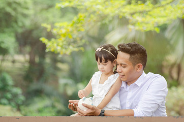 Saving and education concept, young father teach her daughter saving money. Sitting on bench under the tree