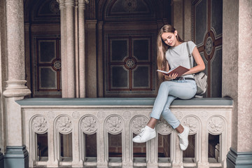 Beautiful and confident. Young female student sitting on the railing in the university hall and reading book.
