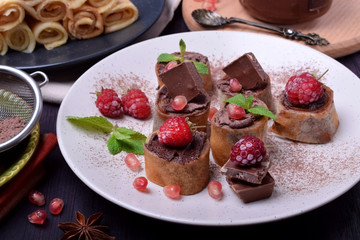 Rolled crepes with chocolate paste topped with raspberries, pomegranate, mint and chocolate pieces on a white plate