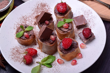 Rolled crepes with chocolate paste topped with raspberries, pomegranate, mint and chocolate pieces on a white plate