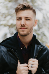Portrait of Attractive Trendy Modern Young Male Model Smiling and Enjoying the Outdoors and the White Winter Snow in the Tree Canopy While Posing in Fashionable Leather Bomber Jacket