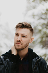 Portrait of Attractive Trendy Modern Young Male Model Smiling and Enjoying the Outdoors and the White Winter Snow in the Tree Canopy While Posing in Fashionable Leather Bomber Jacket