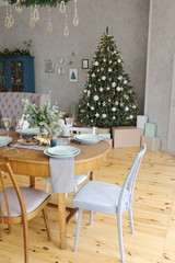 Elegant Christmas tree with gifts in the living room with dinner table near the textured gray walls in luxury apartments. 