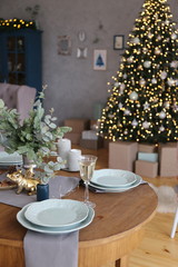Elegant Christmas tree with gifts in the living room with dinner table near the textured gray walls in luxury apartments. 