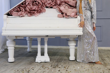white grand piano decorated with pink pale yellow peonies made of paper in luxury suites with gray walls and a silver lamp and barefoot girl in silver dress