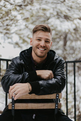 Fototapeta premium Portrait of Attractive Trendy Modern Young Male Model Sitting While Smiling and Enjoying the Outdoors and the White Winter Snow in the Tree Canopy While Posing in Fashionable Leather Bomber Jacket