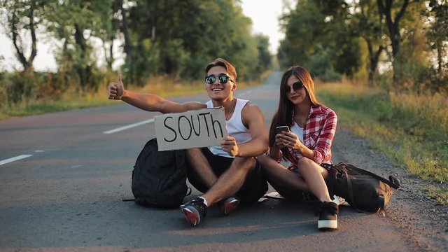 A young couple are hitchhiking sitting on the road and using smartphone. A man and a woman stop the car on the highway with a sign South