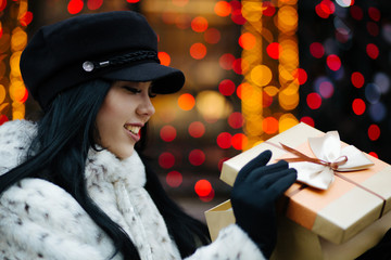 Pretty smiling brunette lady wearing cap and fur coat, holding gift box at the city. Space for text