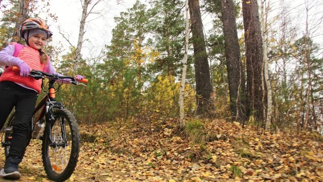 One caucasian children walk with bike in autumn park. Little girl walking black orange cycle in forest. Kid goes do bicycle sports. Biker motion ride with backpack and helmet. Mountain bike hardtail.