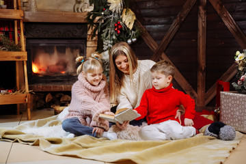 Merry Christmas and Happy New Year. Beautiful family in Xmas interior. Pretty young mother reading a book to her daughter and son near Christmas tree.