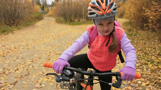 One caucasian children rides bike road in autumn park. Little girl riding black orange cycle in forest. Kid goes do bicycle sports. Biker motion ride with backpack and helmet. Mountain bike hardtail.