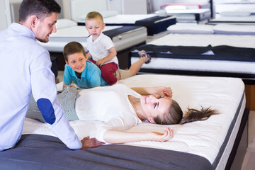 Young cheerful woman with husband and sons lying on back on tested mattress in store