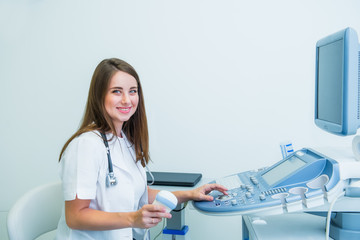 Portrait of young smiling doctor, ultrasound specialist looking at camera and using Ultrasound Scanning Machine for pacient testing. Copy space. Selective focus. - obrazy, fototapety, plakaty