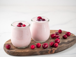 Yogurt with cranberry. Two glass jar with pink yoghurt or milkshake and red cranberries on wooden cutting board over white marble background. Copy space for text