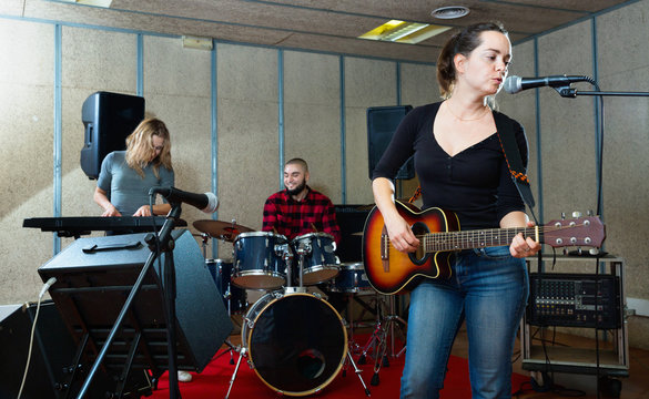 Attractive female soloist playing guitar and singing with her music band in sound studio