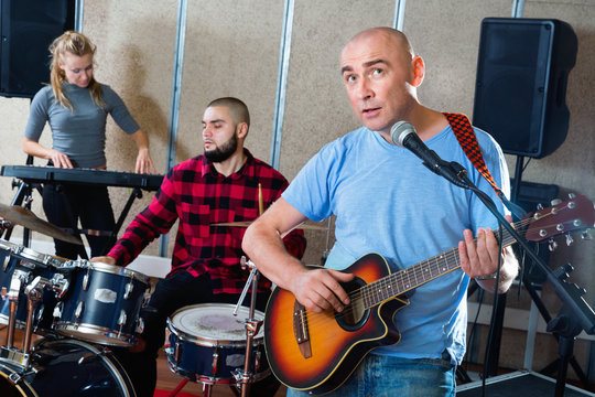 Rehearsal of music group. Band leader playing guitar and singing with his musicians in recording studio