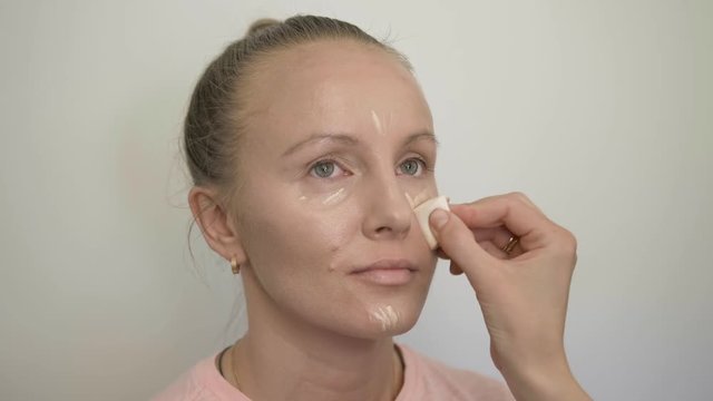 Professional makeup artist applying tone by cosmetic brush for masking eyebags on adult blonde woman model face