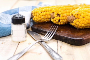 Fresh cooked corn on cutting board  on rustic wooden table, knife and fork, salt 