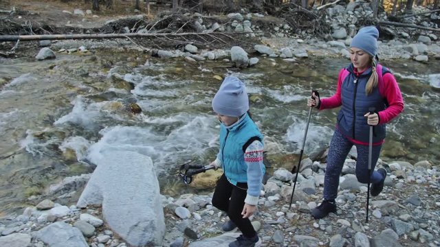 Woman do Nordic walking in nature near mountain river. Girls and children use trekking sticks and nordic poles, backpacks. Family travels sports. Kid is learning from mother and grandmother the proper