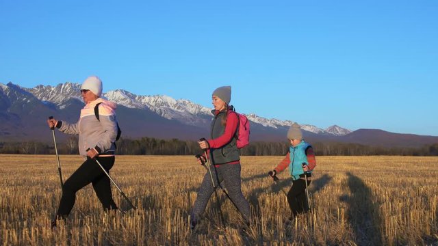 Woman Nordic walking in nature on background of mountains in field. Girls and children use trekking sticks and nordic poles, backpacks. Family travels sports. Kid is learning from mother and