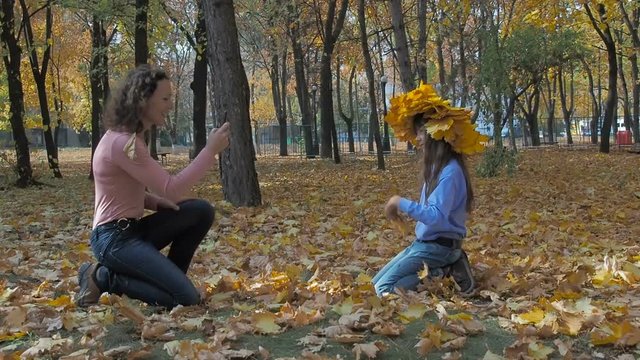 A woman on a smartphone photographs a child. A woman photographs on a smartphone a little girl in a wreath of yellow leaves in an autumn park.