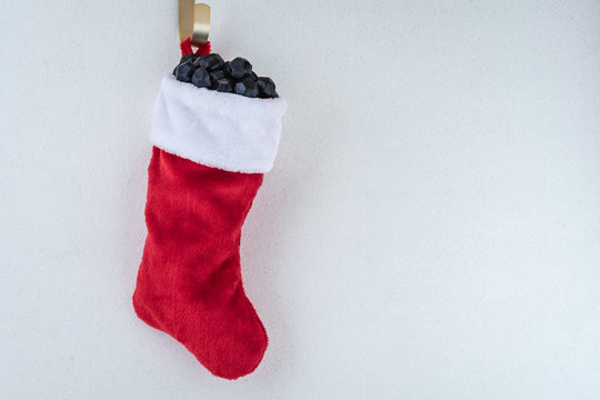 Traditional red and white plush Christmas stocking stuffed with coal shaped candy on a gold hook against a white background with silver flecks, as a naughty for Christmas concept background