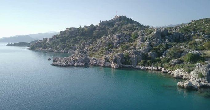 Forward aerial drone footage over water to coast of Simena Kalekoy Village over rocky outcrop to reveal boats anchored in natural cove in Kekova Island, Ucagiz, Turkey. 4k at 23.97fps
