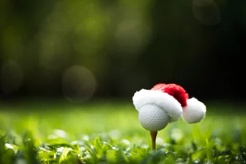 Schilderijen op glas Festive-looking golf ball on tee with Santa Claus' hat on top for holiday season on golf course background © amenic181