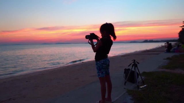 Silhouette little girl using camera taking photo on beach in sunset with twilight beautiful sky, slow-motion