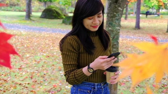 Happy Woman Using Smartphone in Park in Autumn Season. Standing under the Tree