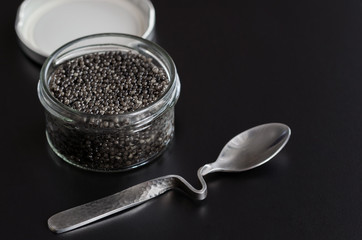 Black caviar in glass jar with spoon on black background with copy space.
