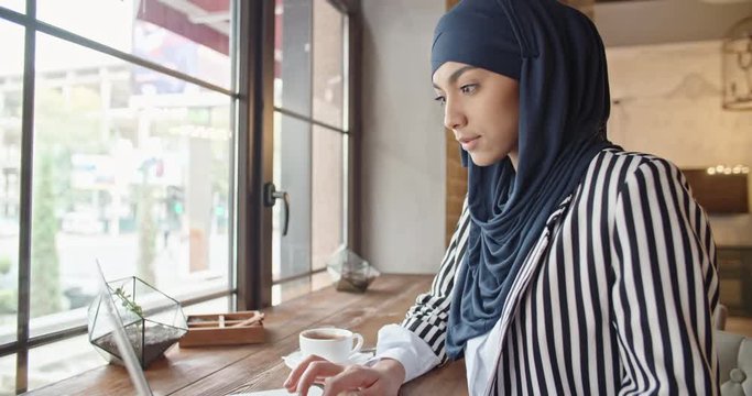 Attractive muslim woman in hijab working on project at laptop, sitting in loft cafeteria, searching in web or studying - closeup 4k