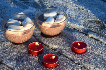 Obraz na płótnie Canvas Two cups of cocoa and marshmallows stand on a wooden table with hoarfrost. Three red candles are burning. Hot drink and marshmallows.