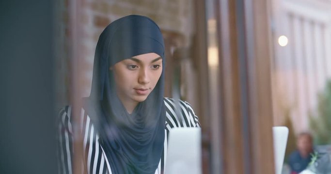 Young muslim woman wearing hijab sitting in modern cafe, working, searching or studying at her laptop, drinking morning coffee - modern muslim concept closeup 4k